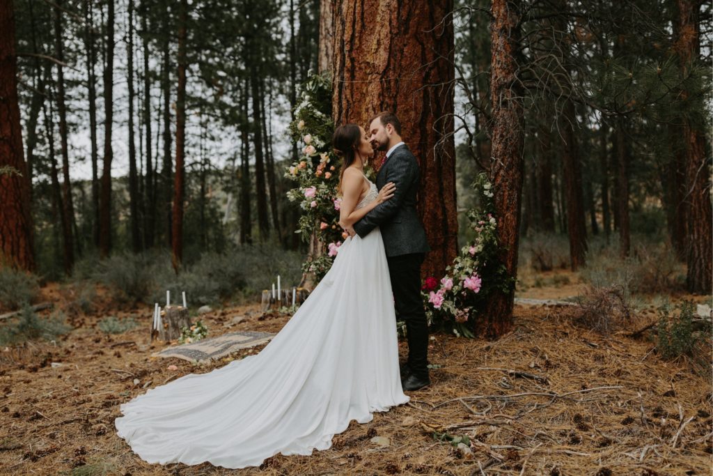 bride and groom embracing in the forest for their woodsy intimate elopement wedding with wild and romantic floral installation on the tree