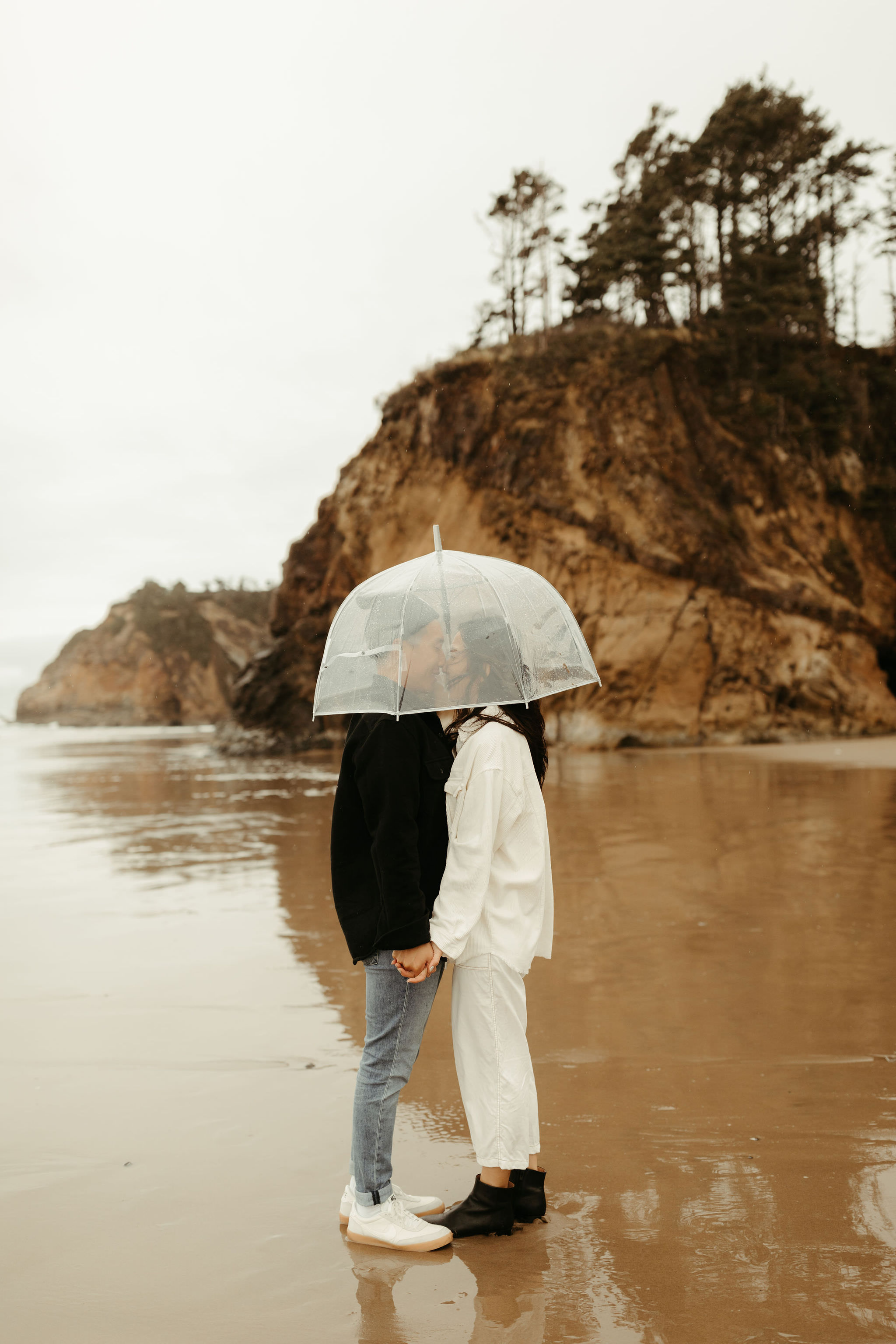 couple at Cannon Beach on a rainy day with a clear umbrella for their moody engagement photos